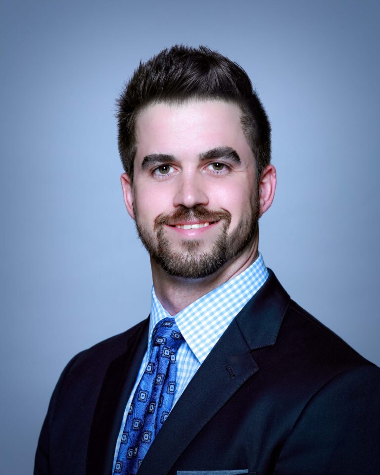 Finished headshot of a smiling man in a navy suit and blue patterned tie on a light blue backdrop